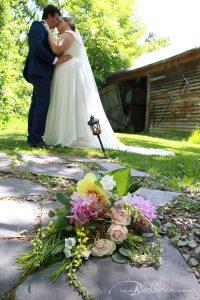 Gorgeous Bridal Bouquet in front of rustic barn in Saratoga NY with Bride and Groom Kissing in background.